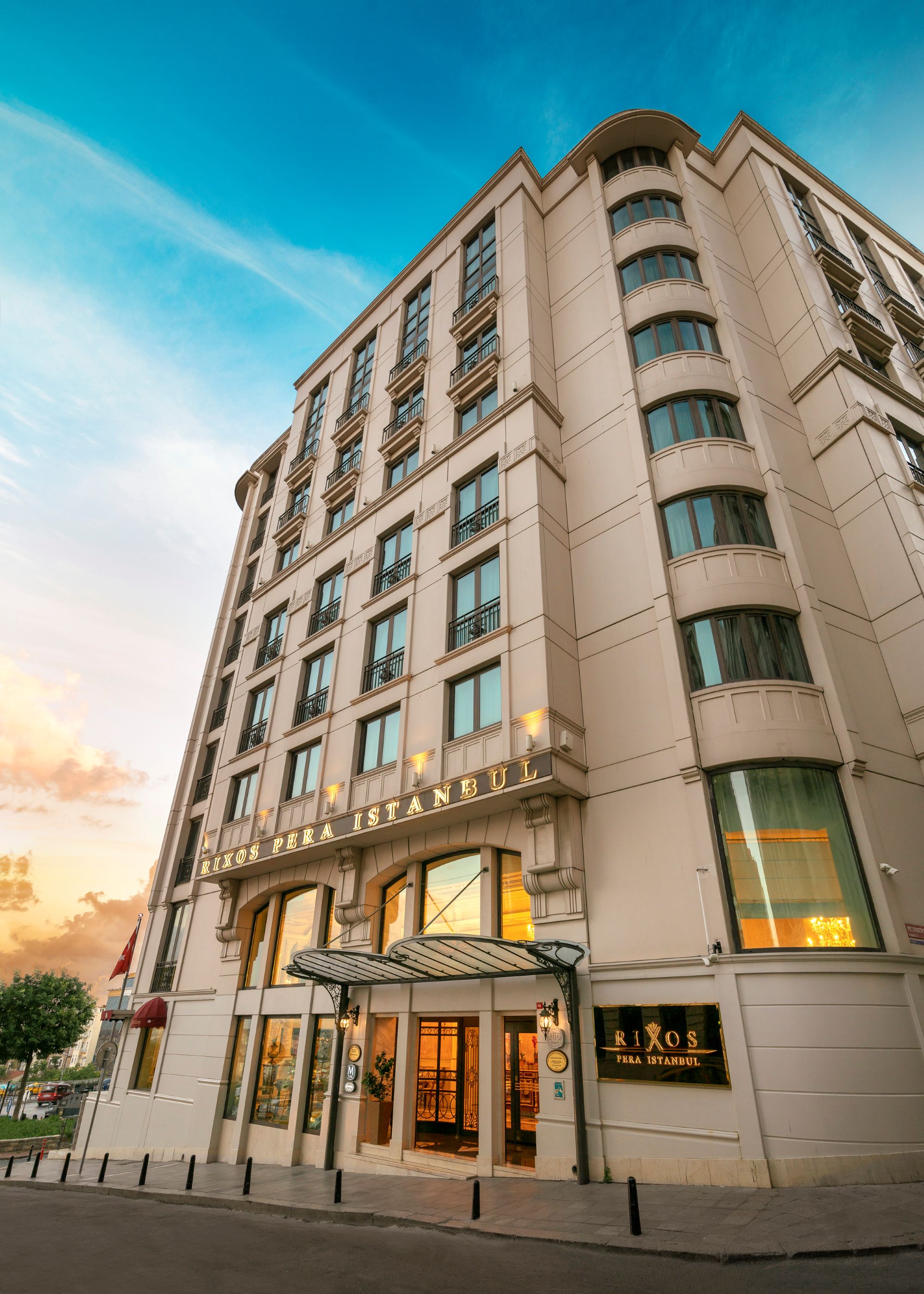 A dramatic, upward-angle photograph of the exterior of the Rixos Pera Istanbul hotel. The tall, traditional stone building features several rows of windows and a covered main entrance at the bottom, all set against a bright blue and cloudy sky.