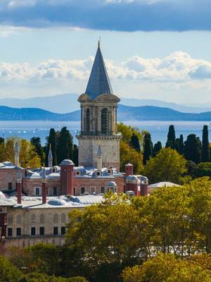 View of Istanbul's historic Topkapı Palace, with its domes and conical tower, framed by trees against the sea and mountains.